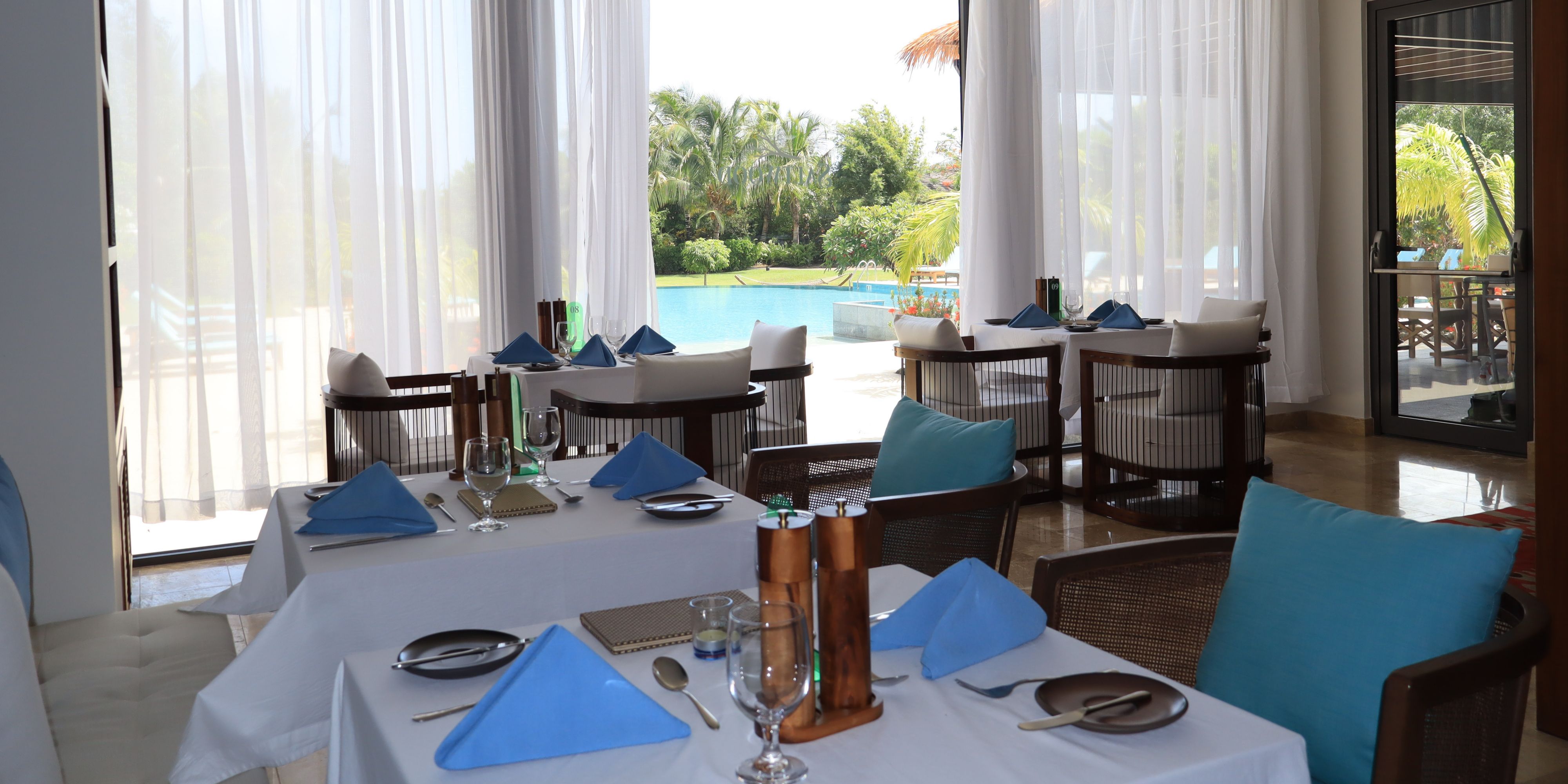 Elegant dining tables set with blue napkins and white linens at Saltwood restaurant, with floor-to-ceiling windows overlooking a tropical pool and lush gardens.