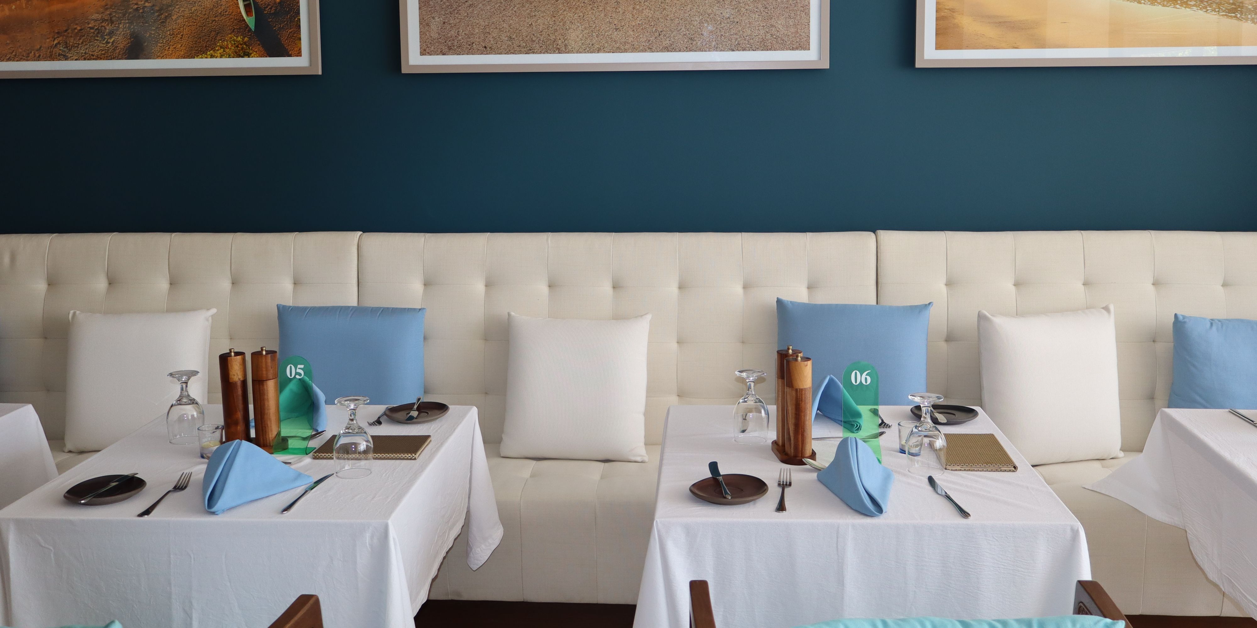 Banquette seating at Saltwood restaurant with tables dressed in white linens and blue accents beneath framed beach-inspired artwork and a deep blue feature wall.