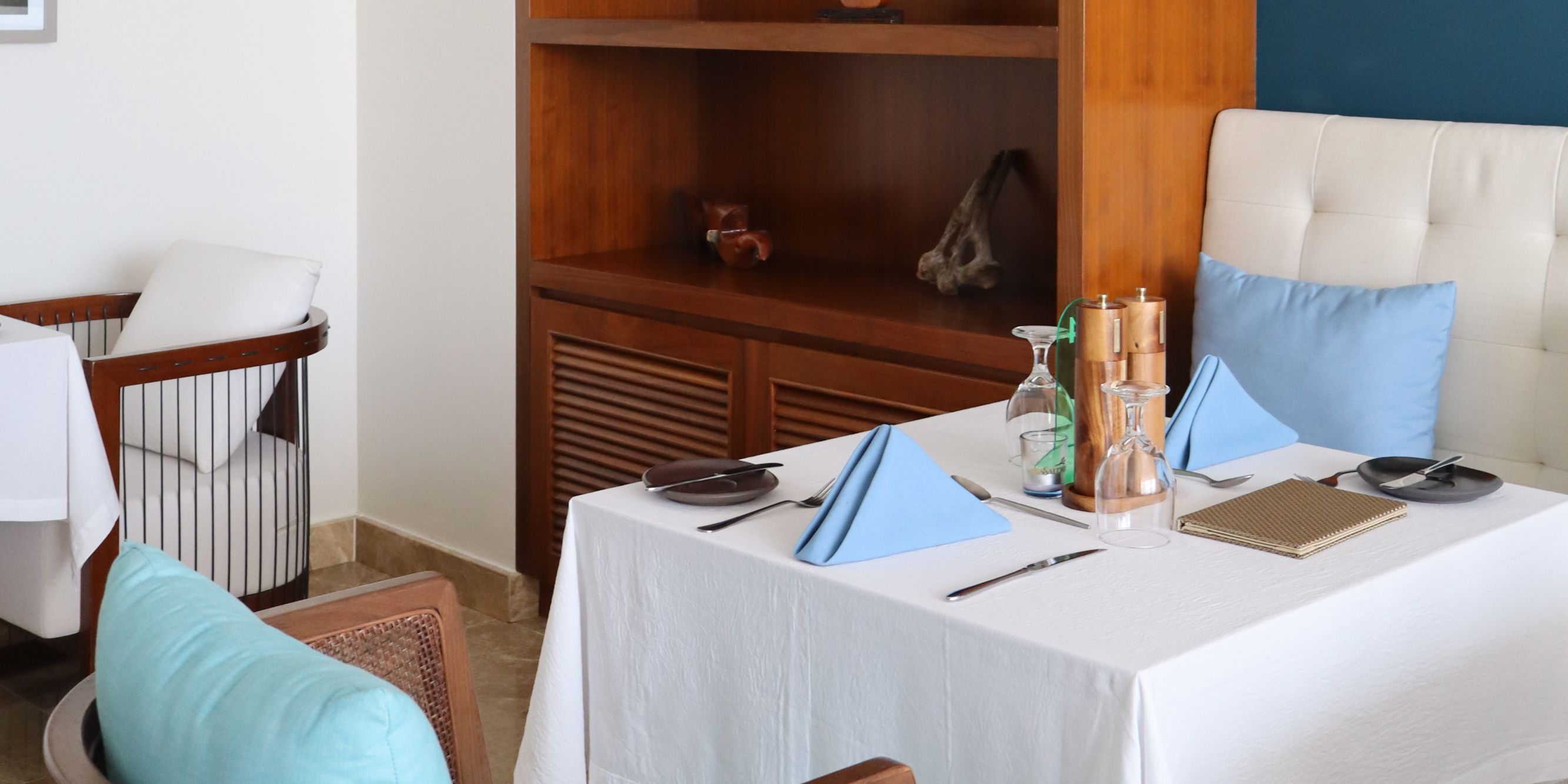Saltwood restaurant dining area featuring a table set with white linens and blue napkins, framed by warm wood shelving, coastal artwork, and comfortable upholstered seating.
