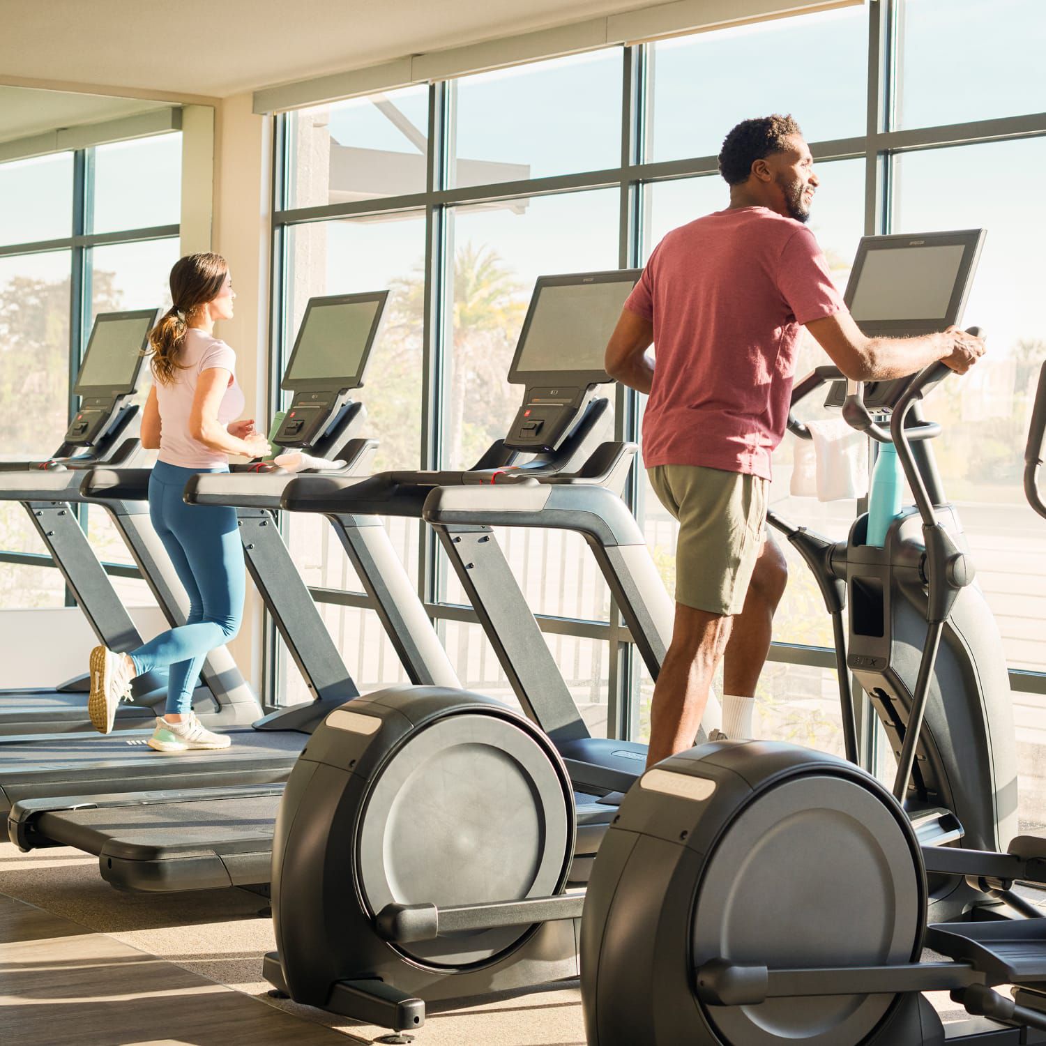 A man working out on an elliptical and a woman running on a treadmill in front of floor-to-ceiling windows.