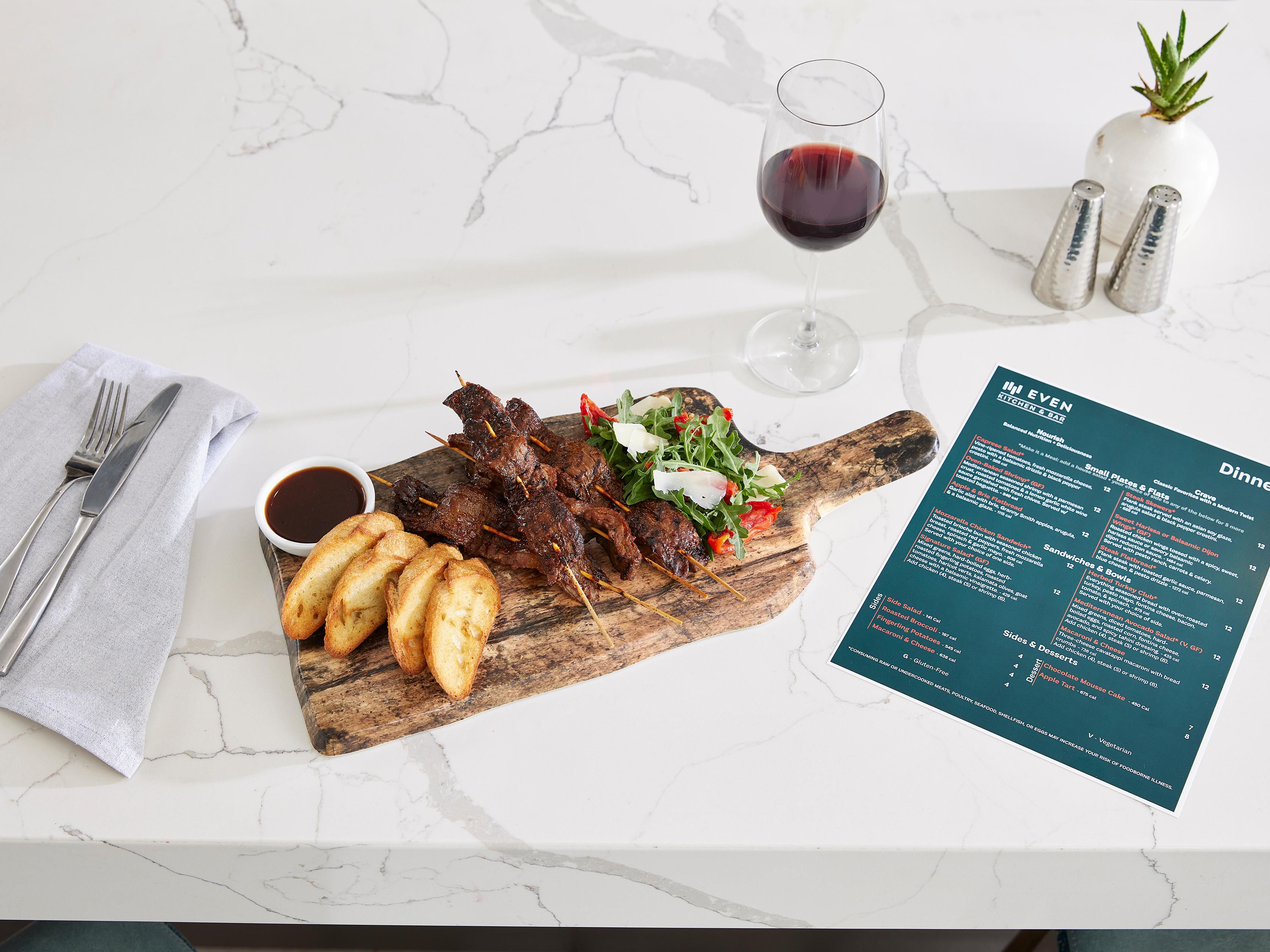 A cutting board with steak skewers, potato wedges, and a salad