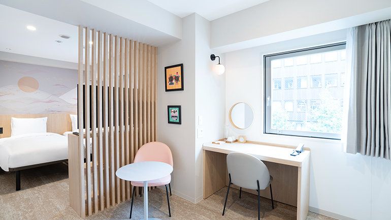 Bright hotel room with a wooden slat divider, small table and chairs, and a desk by a window.