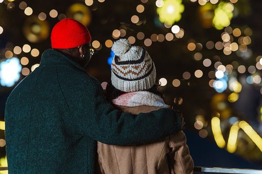 A couple embraces while enjoying the warm glow of festive lights in a winter setting. The intimate scene captures the essence of warmth and togetherness during the holiday season.