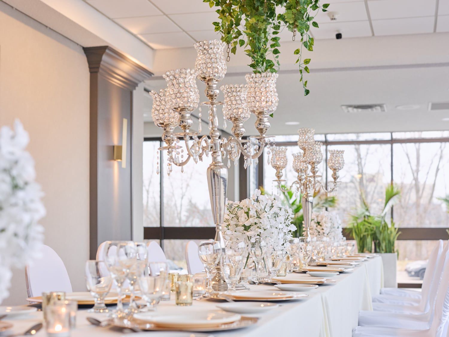 Elegant wedding reception table set with flowers, glassware, and chandeliers in a bright event room.