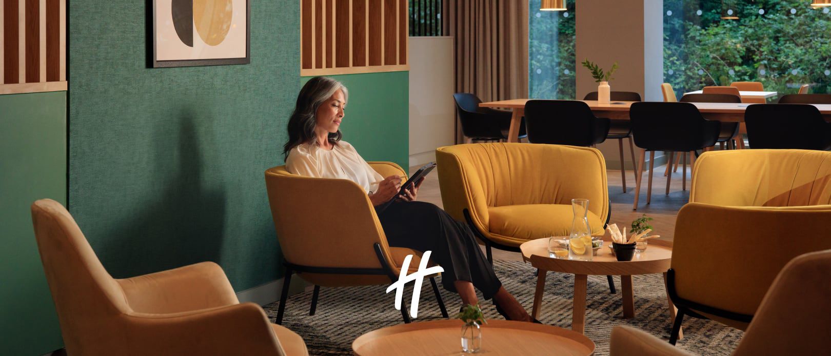 Guest sitting in a modern yellow lounge chair in a modern hotel lobby with green walls, wood accents, and warm seating.