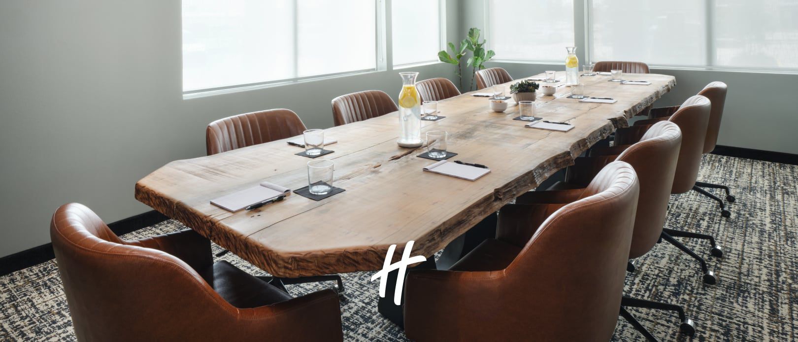 Large wooden conference table set with notebooks and water glasses in a bright meeting room with leather chairs.
