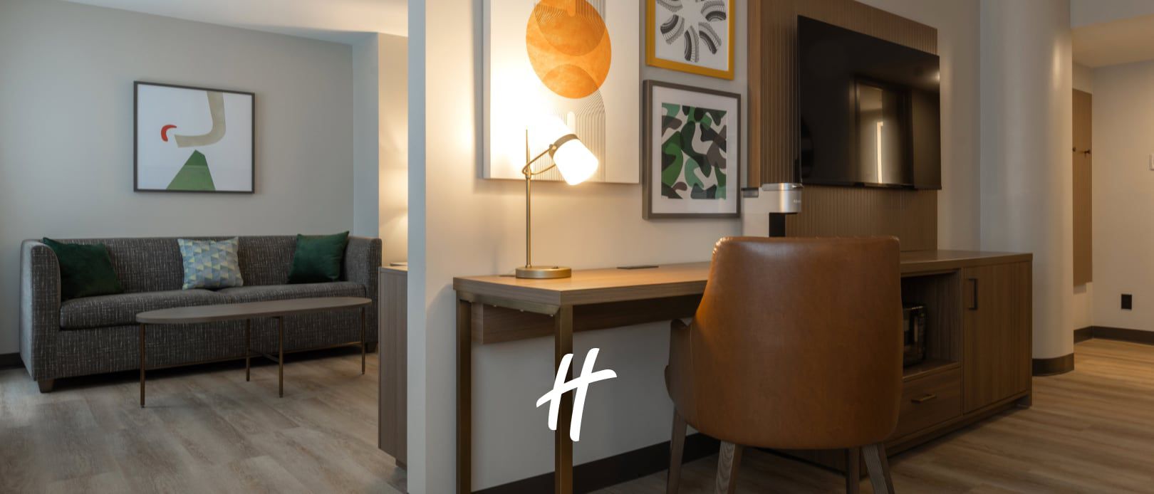 Modern hotel guest room with a sofa, desk, chair, artwork, and warm bedside lighting.