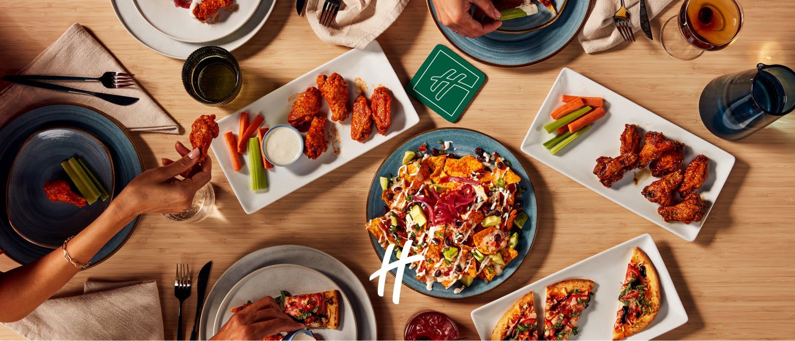 Overhead view of a restaurant table with pizza, wings, salad, drinks, and hands reaching for food.