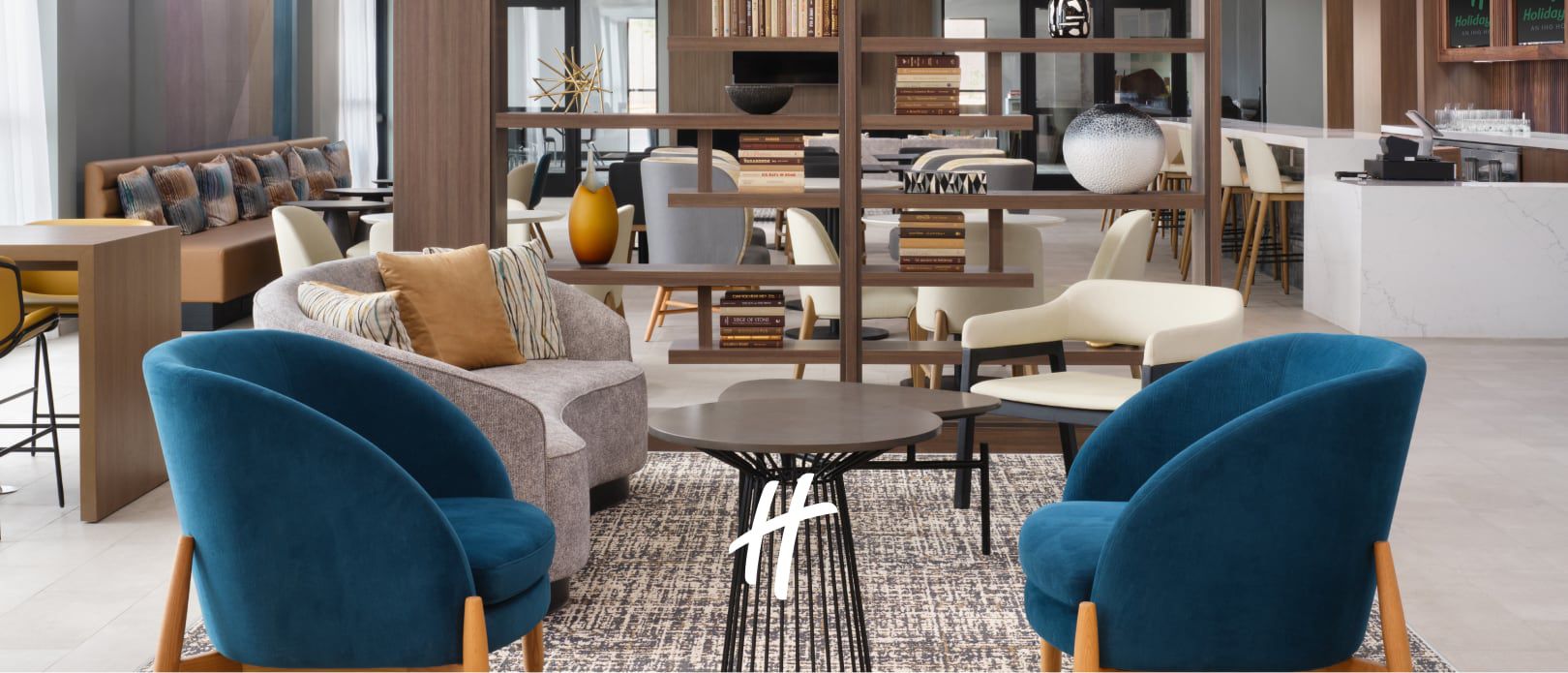 Modern hotel lounge with blue chairs, a sofa, coffee tables, and an open kitchen area in the background.