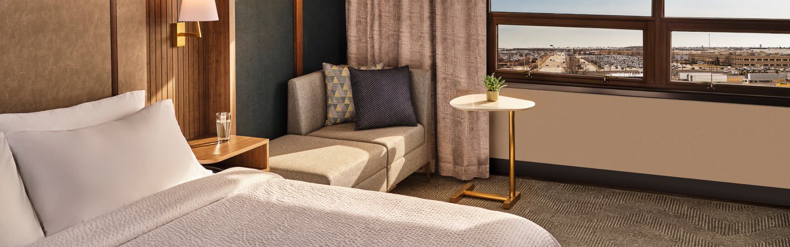 Modern hotel guest room with a bed, window seat, side table, and city view.