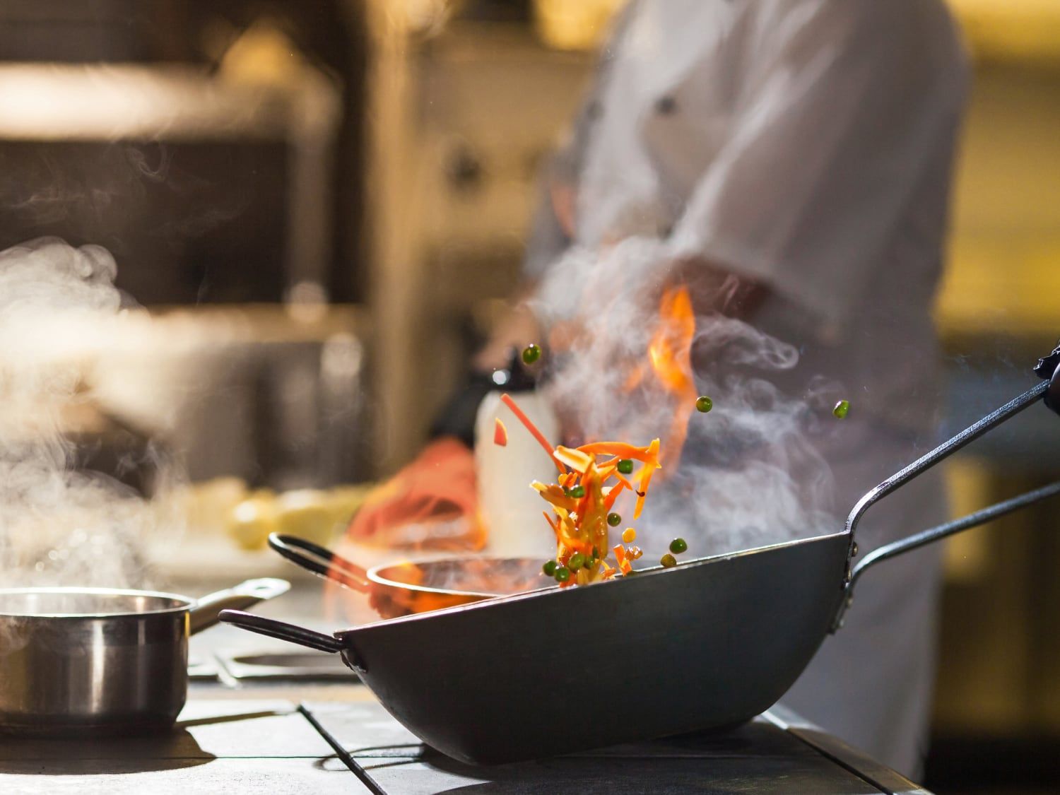 A chef cooking food in a wok with veggies and flames flying out of the pan