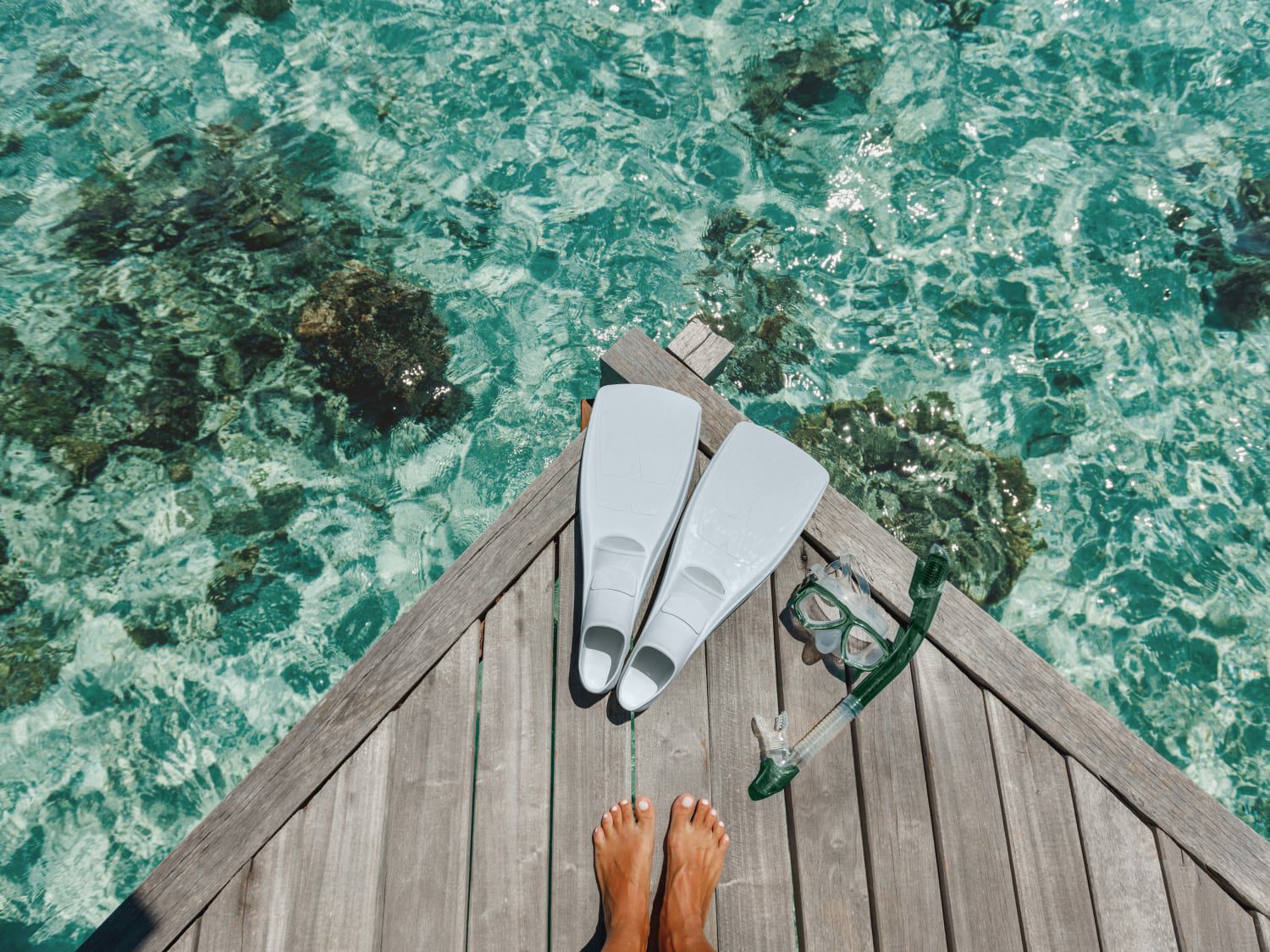 A pair of feet standing on a dock with a snorkel mask and flippers overlooking crystal clear water