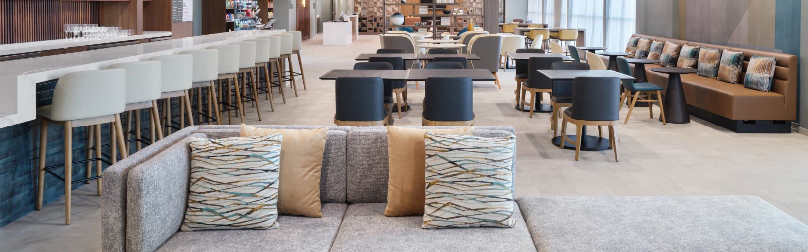 Modern hotel lobby with a gray sofa, patterned pillows, bar seating, dining tables, and lounge areas.