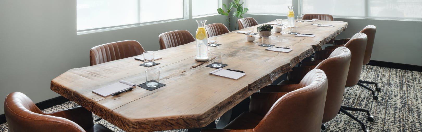 Large wooden conference table set with notebooks and water glasses in a bright meeting room with leather chairs.