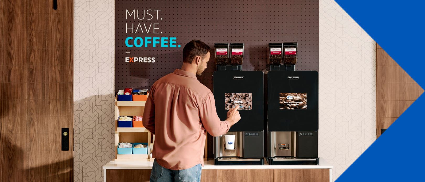A guest uses a bean-to-cup coffee machine in a Holiday Inn Express breakfast area with a “Must. Have. Coffee.” sign on the wall.