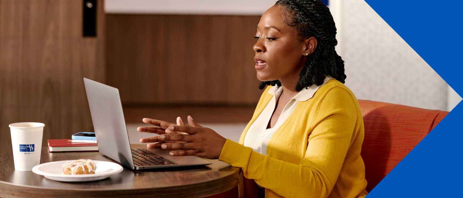 A woman in a yellow sweater works on a laptop at a table with coffee and a pastry nearby.
