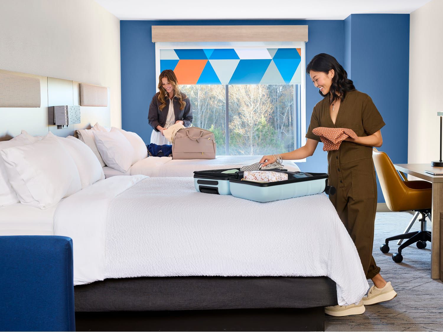 Two women unpacking in a double queen room with a view of greenery out the window.