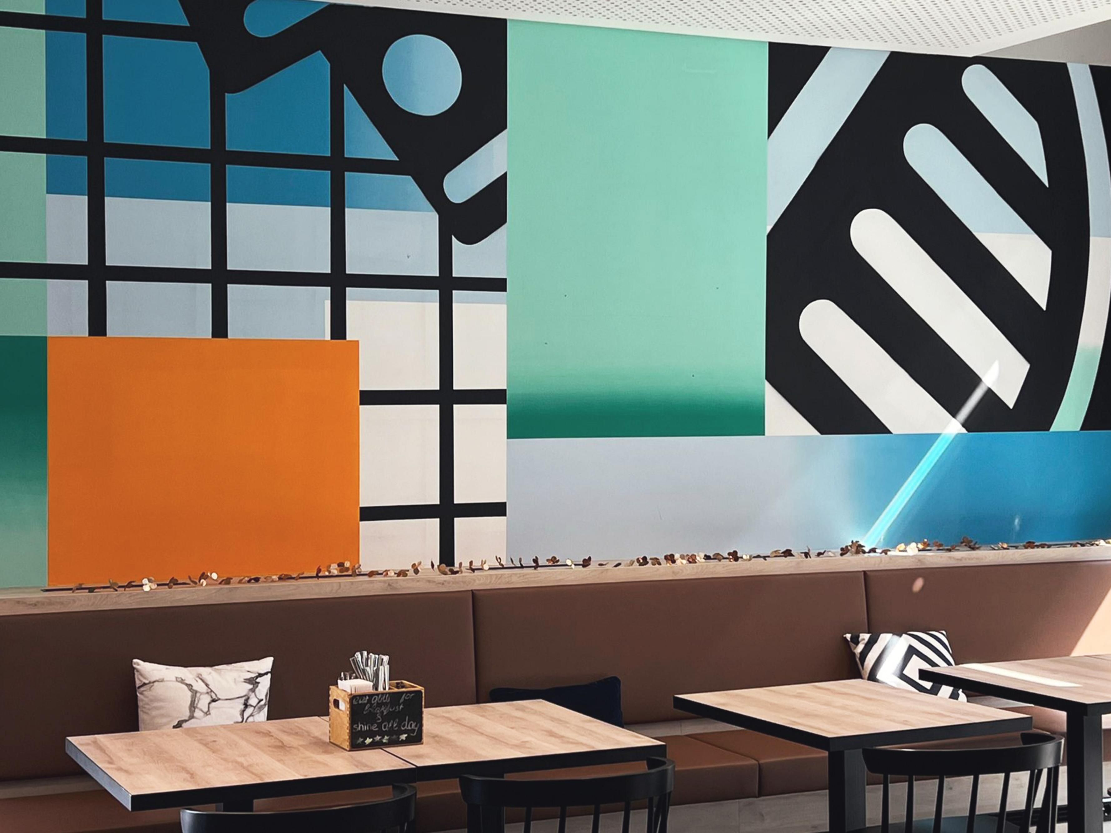 Isakov brings his urban artistry to the niu Kettle in Stuttgart. Shades of green, blue and rich orange dance across the walls, combining architectural motifs with bold ornamentation. A translator, poet and compositor, he turns each mural into a striking visual story—dynamic, playful and utterly unforgettable."