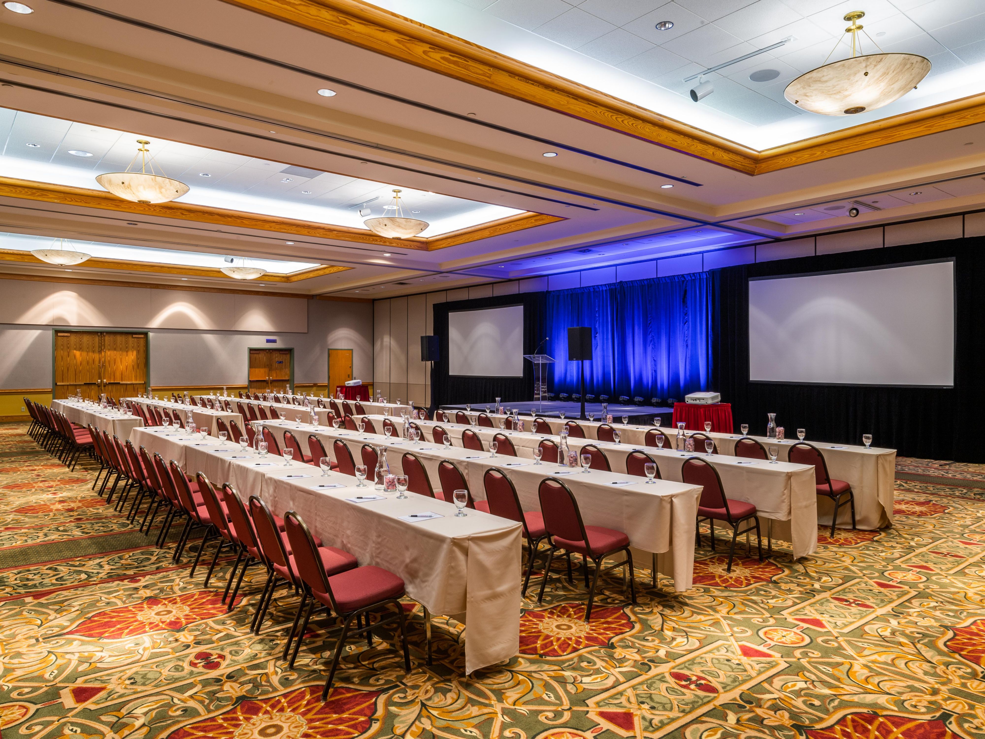 Large meeting room with long tables, stage, and projector screens