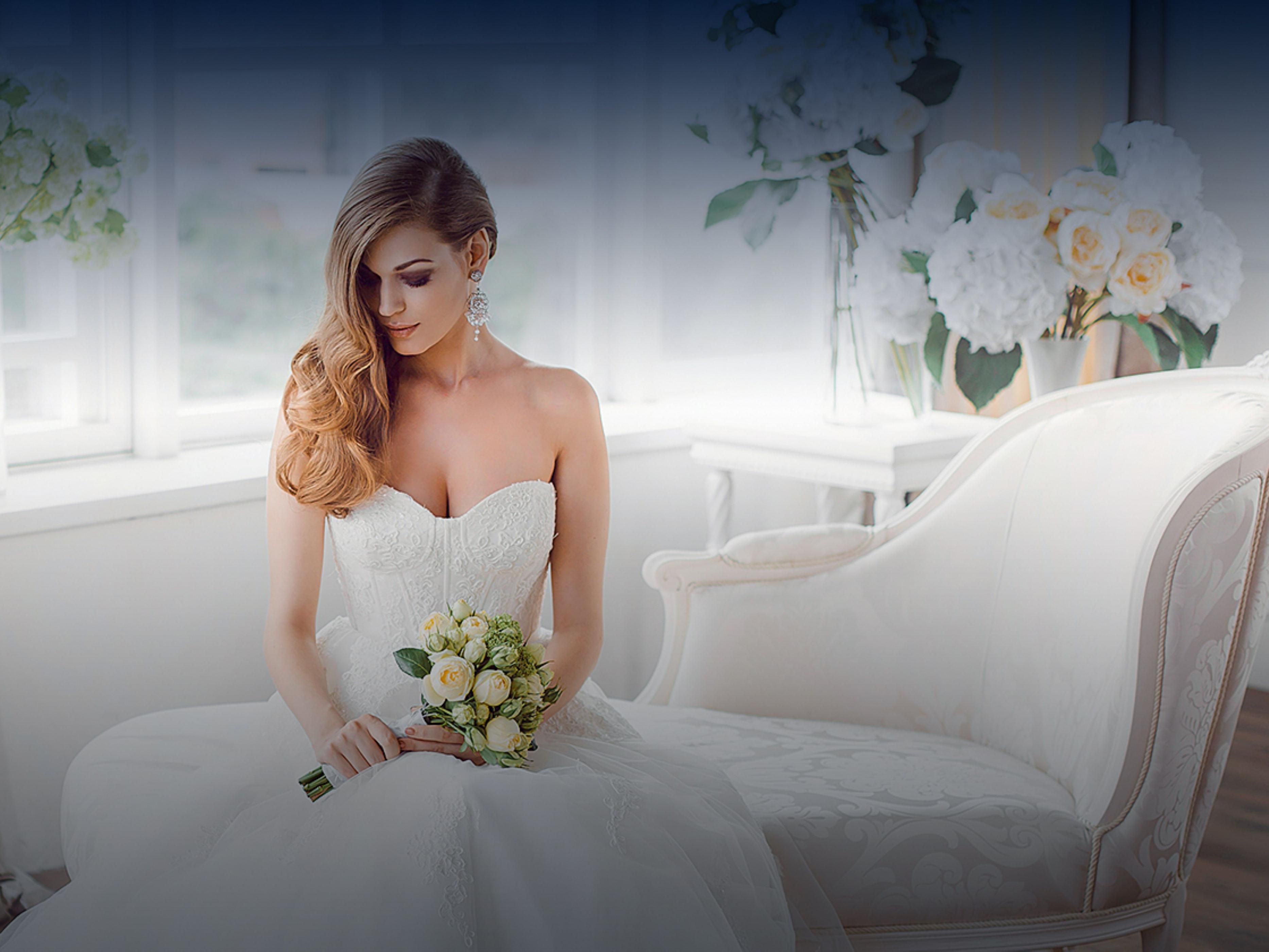 Bride in a white gown sits on an ornate chaise with a bouquet.