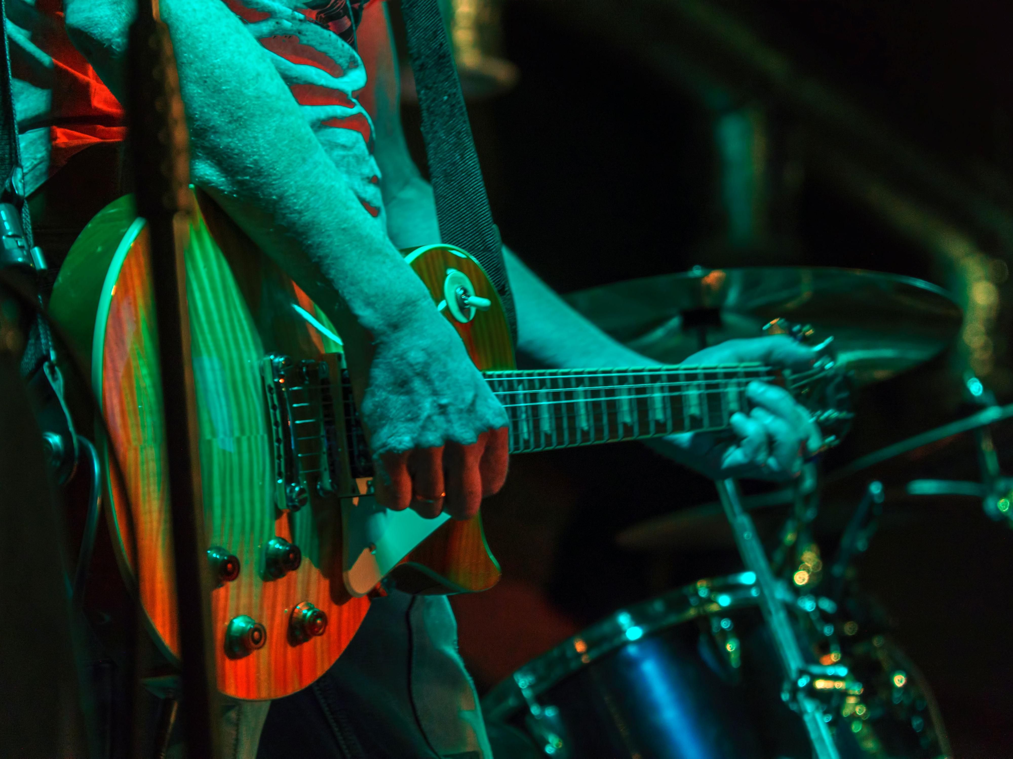 Close-up of a musician playing an electric guitar on stage with soft lighting and a drum set in the background.