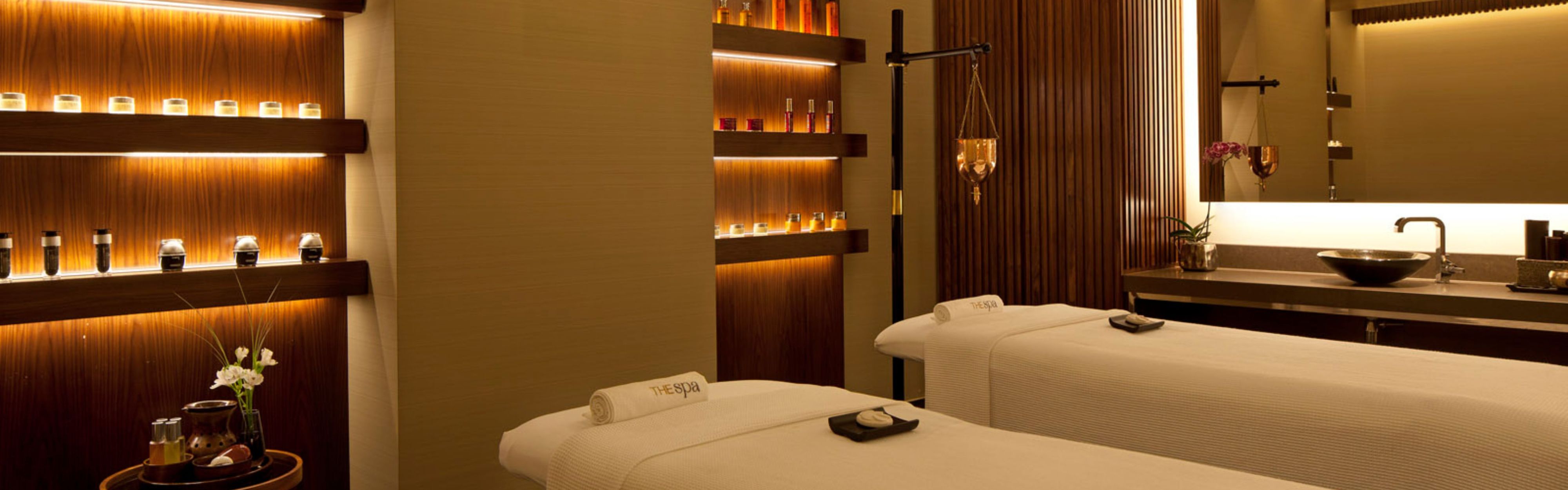 Spa treatment room with two massage tables, warm lighting, and product shelves
