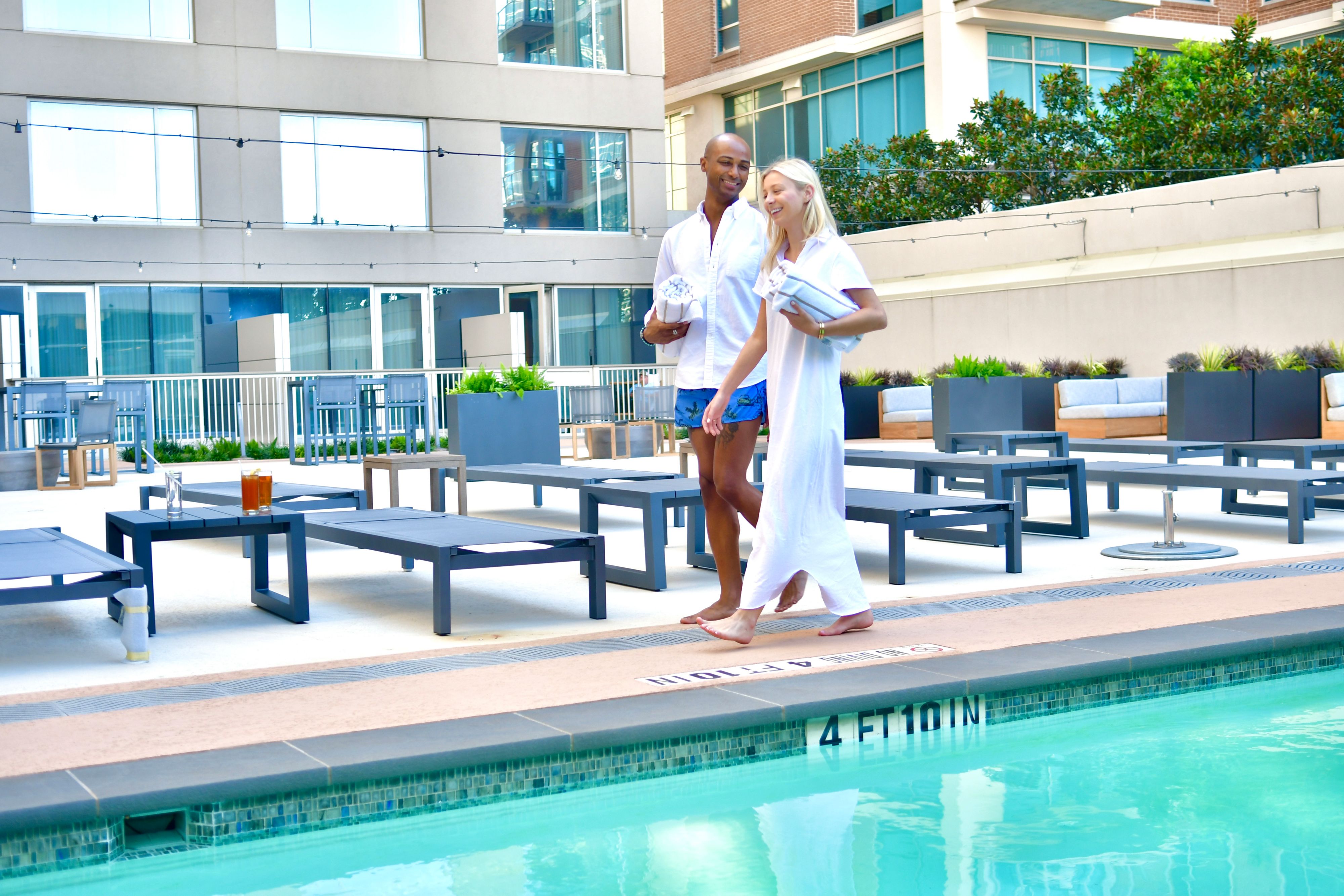 Guest in a white cover-up and another guest carrying towels walk beside an outdoor hotel pool surrounded by lounge chairs and a modern building.