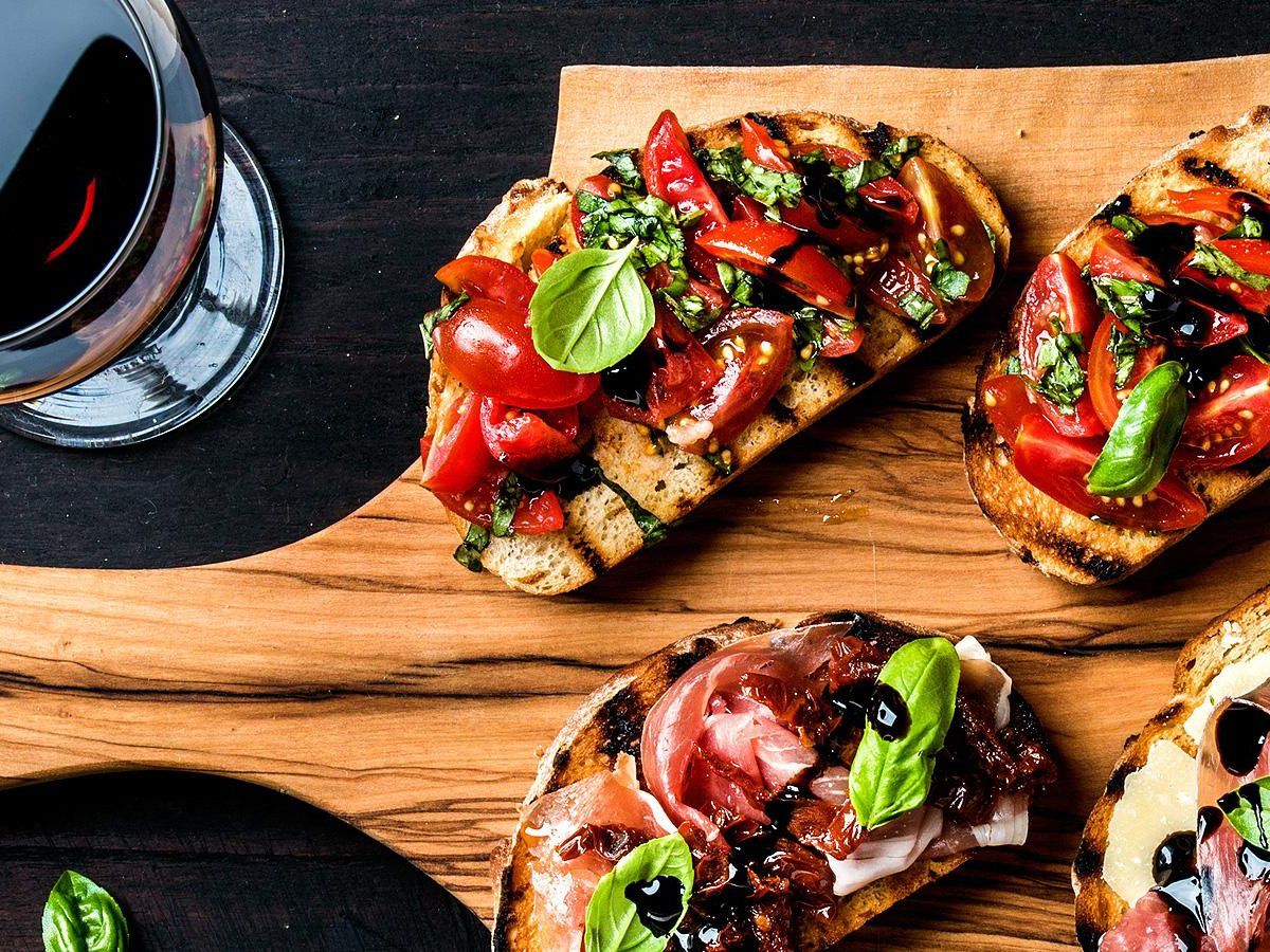 Toasted tartines topped with tomatoes, herbs, and cured meat on a wooden board beside a glass of red wine.