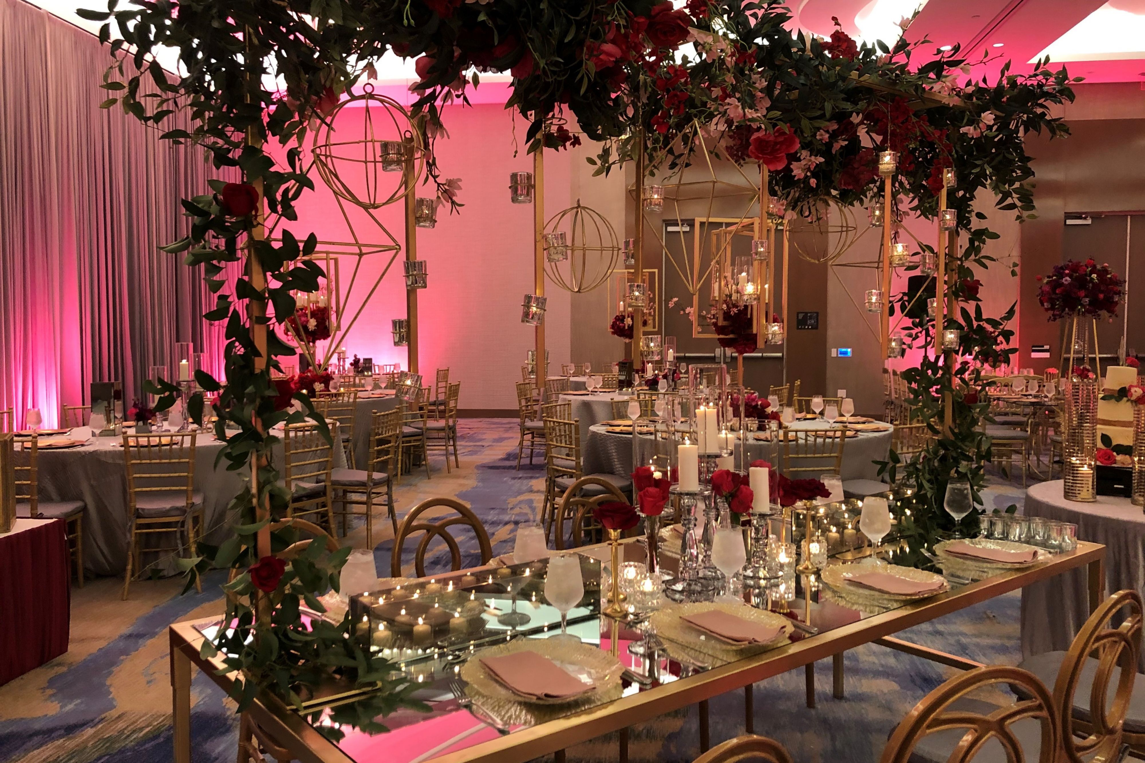 Elegant banquet space featuring a mirrored head table beneath a floral canopy, with candlelit place settings, gold chairs, and pink ambient lighting.