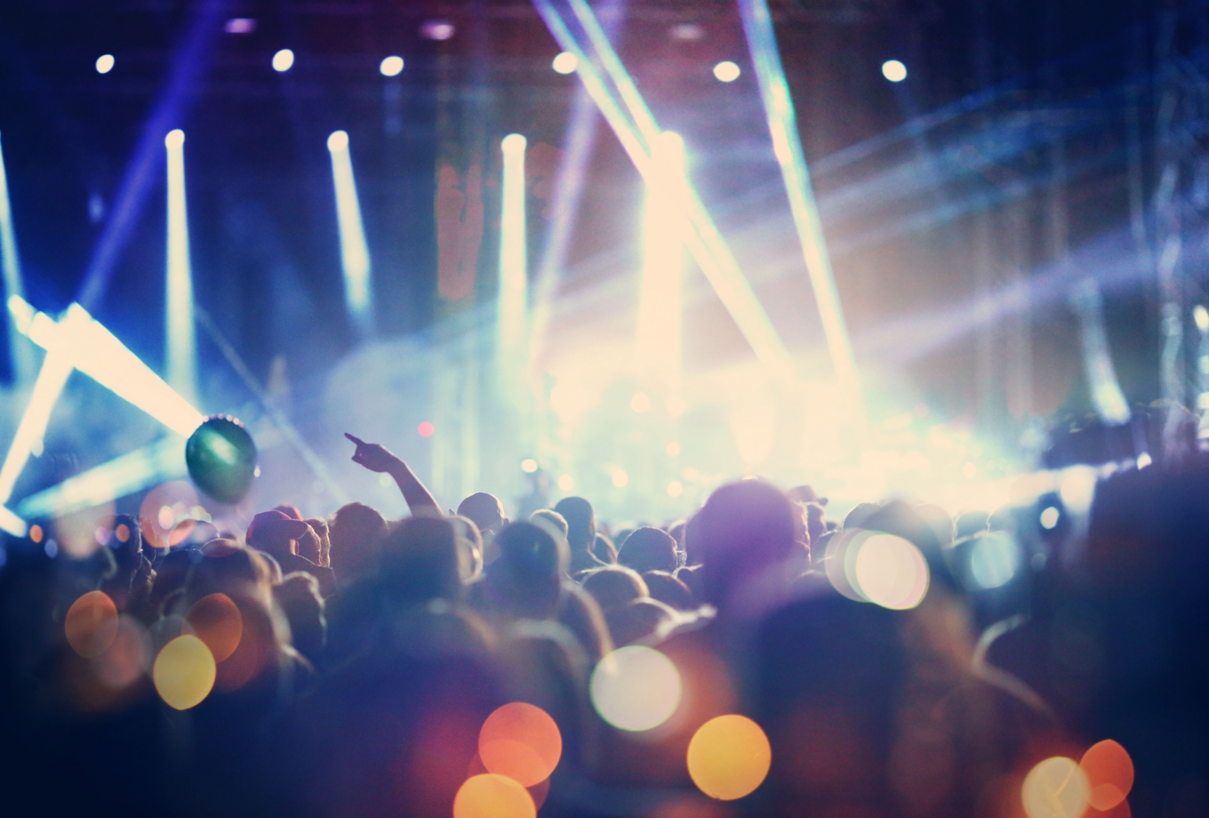 Rear view of people cheering at concert during music festival.Large group of unrecognizable adults with their arms raised.Some of them holding beer cans,some are taping the show with phones.There's a stage and a band performing in background,out of focus.Stage is toned purple blue.
