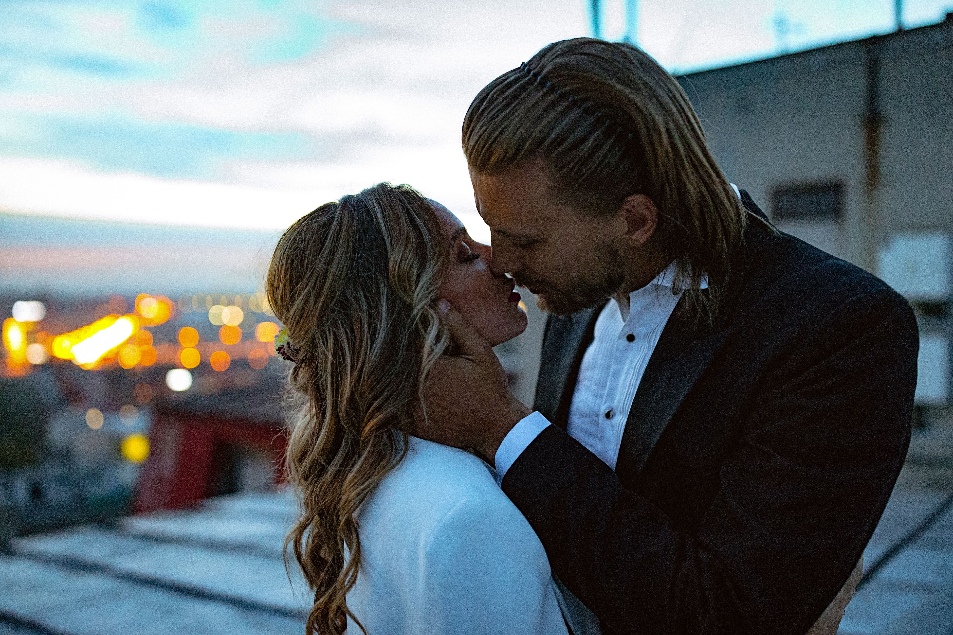 Affectionate bride and groom, embraces and kisses on the rooftop at sunset. Blurred city lights are visible in the background
