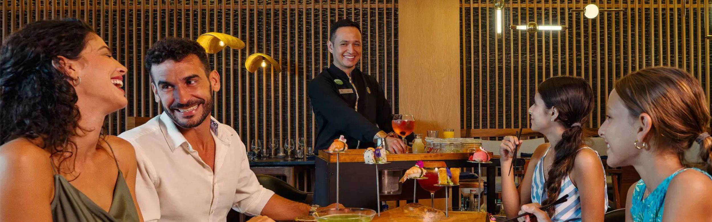 Huéspedes sentados alrededor de una mesa de restaurante disfrutando de alimentos y bebidas mientras un camarero presenta platos de un carrito con ruedas.
