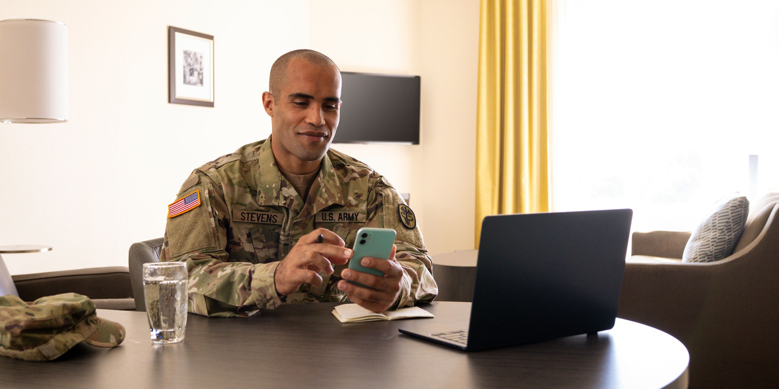 Service member in hotel room looking at mobile phone, with laptop open on work table.