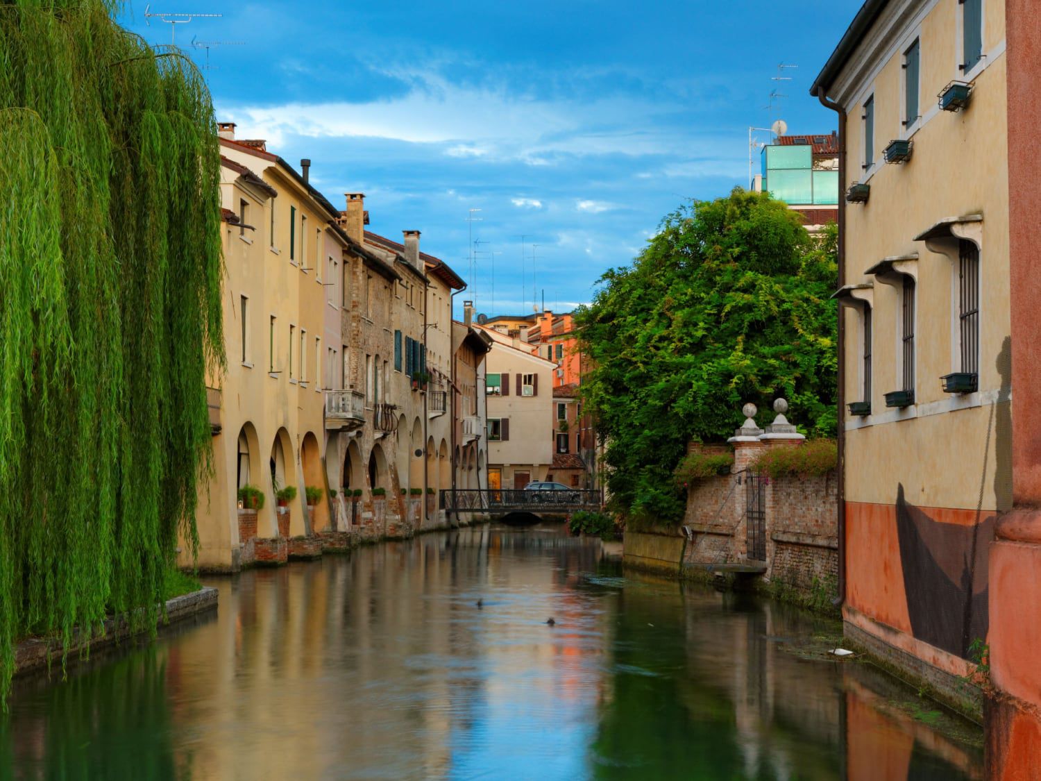 Canal with pastel buildings, greenery, and a distant bridge under a blue sky