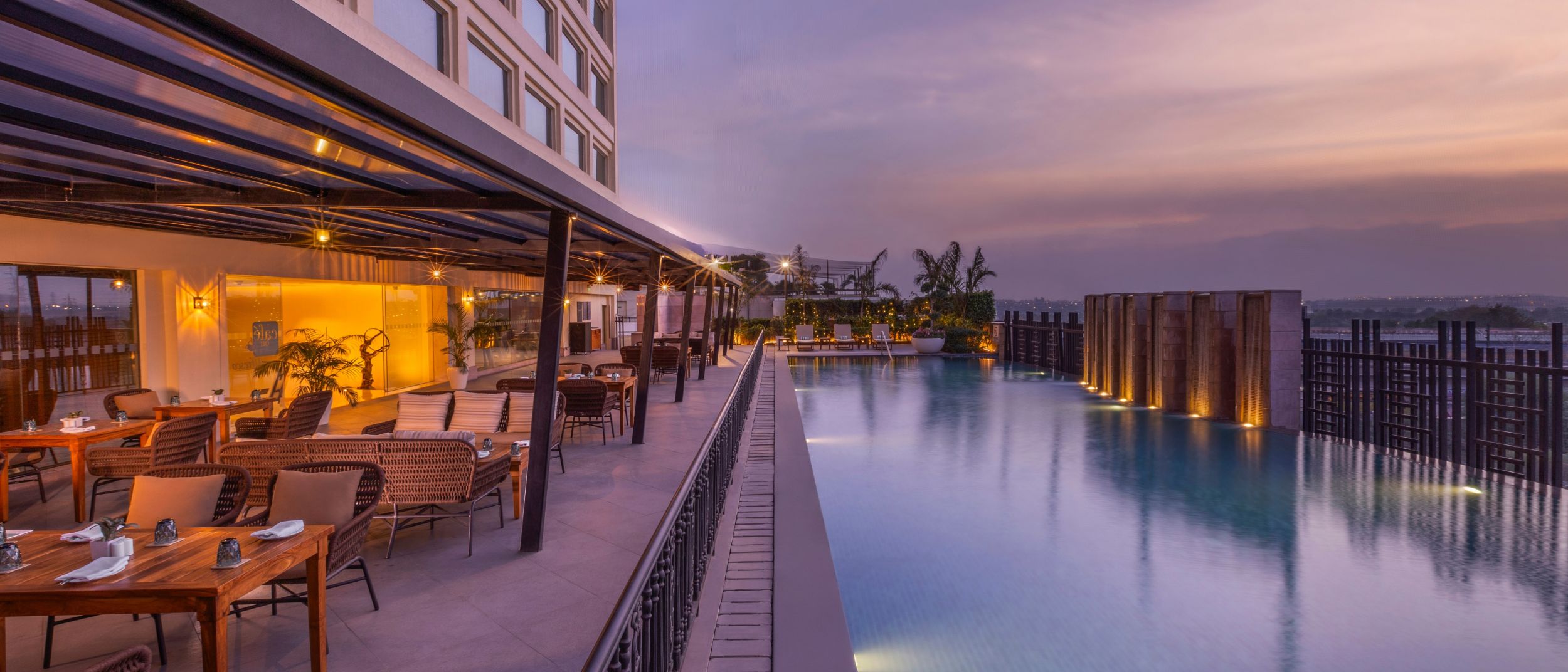 Sunset view of a luxury rooftop infinity pool and warmly lit open-air restaurant overlooking the city.