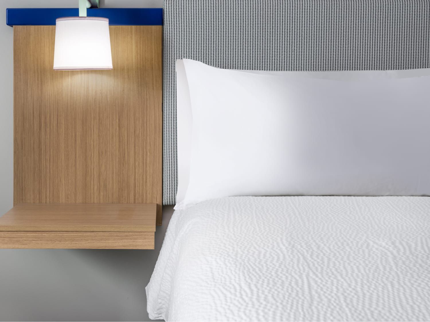 Modern hotel bed with white linens and a wood nightstand with a lamp.
