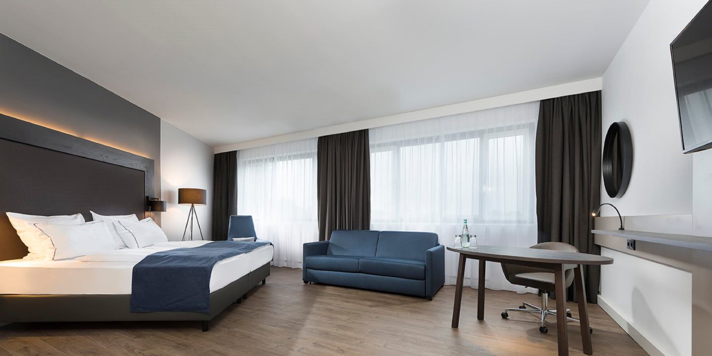 Modern hotel room with king bed, sofa, desk, and large windows.
