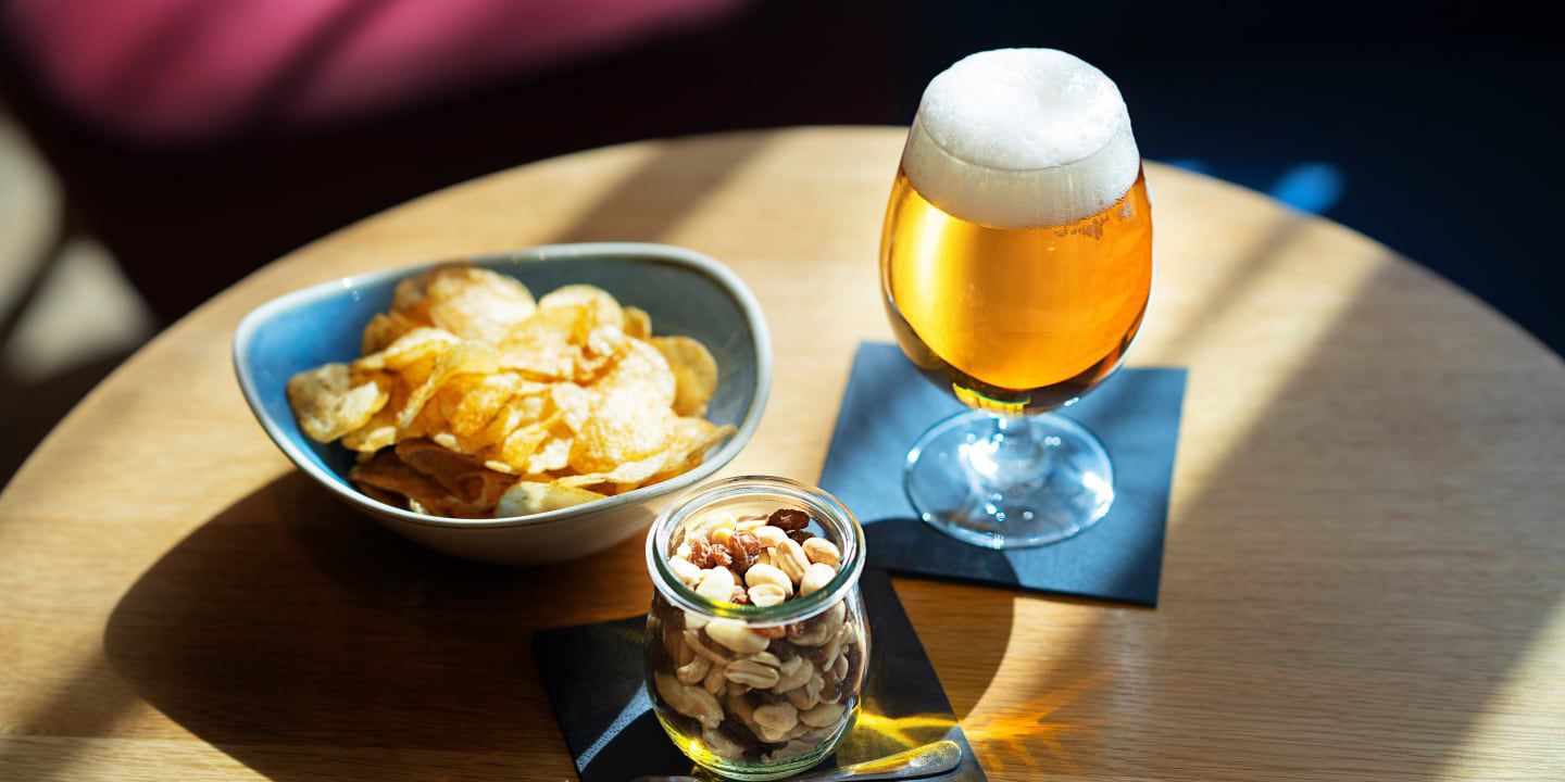 Glass of beer with bowl of chips and jar of mixed nuts.