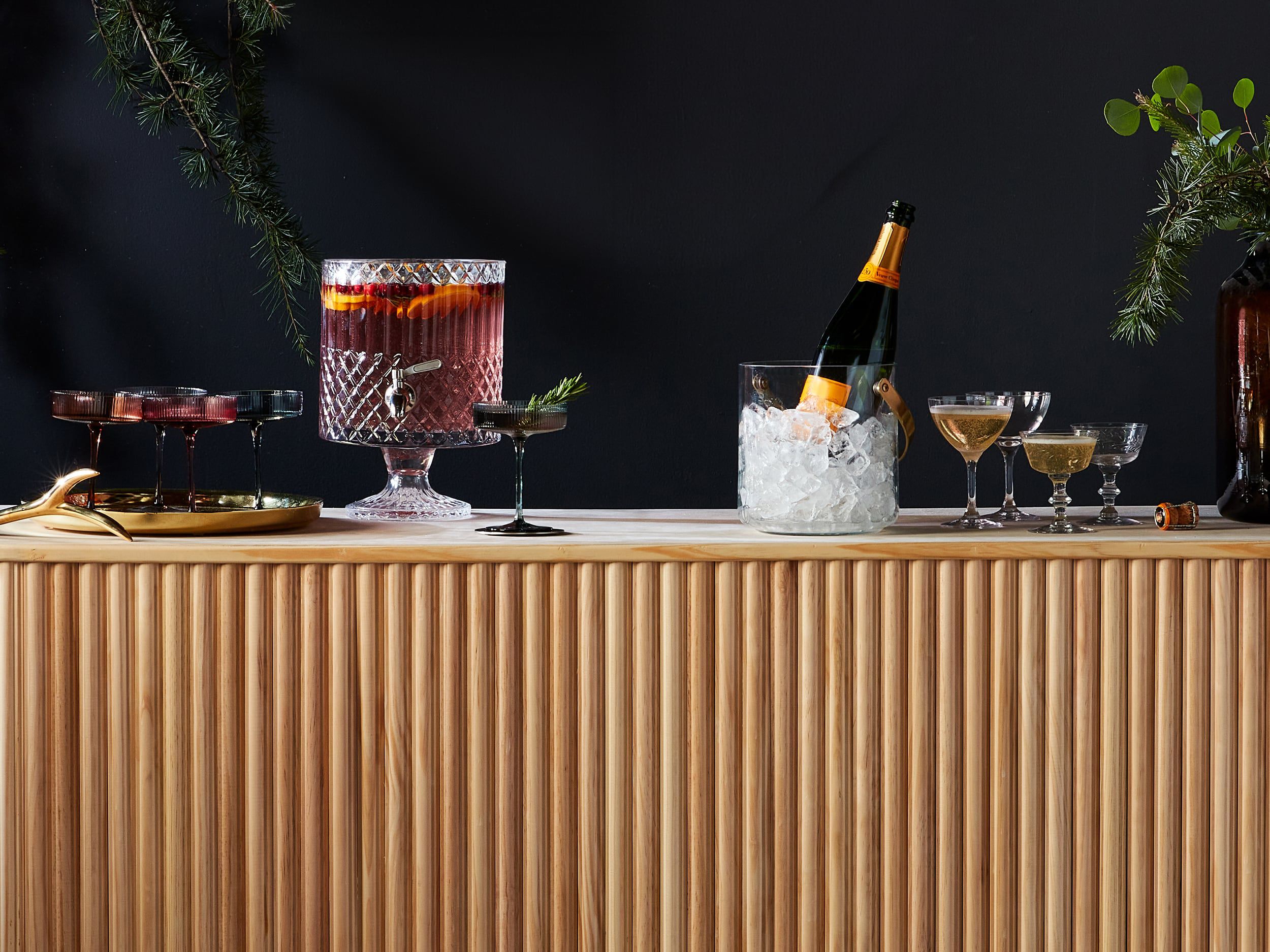 A festive bar setup featuring a wooden counter with a champagne bottle in an ice bucket, coupe glasses filled with drinks, and a punch bowl.