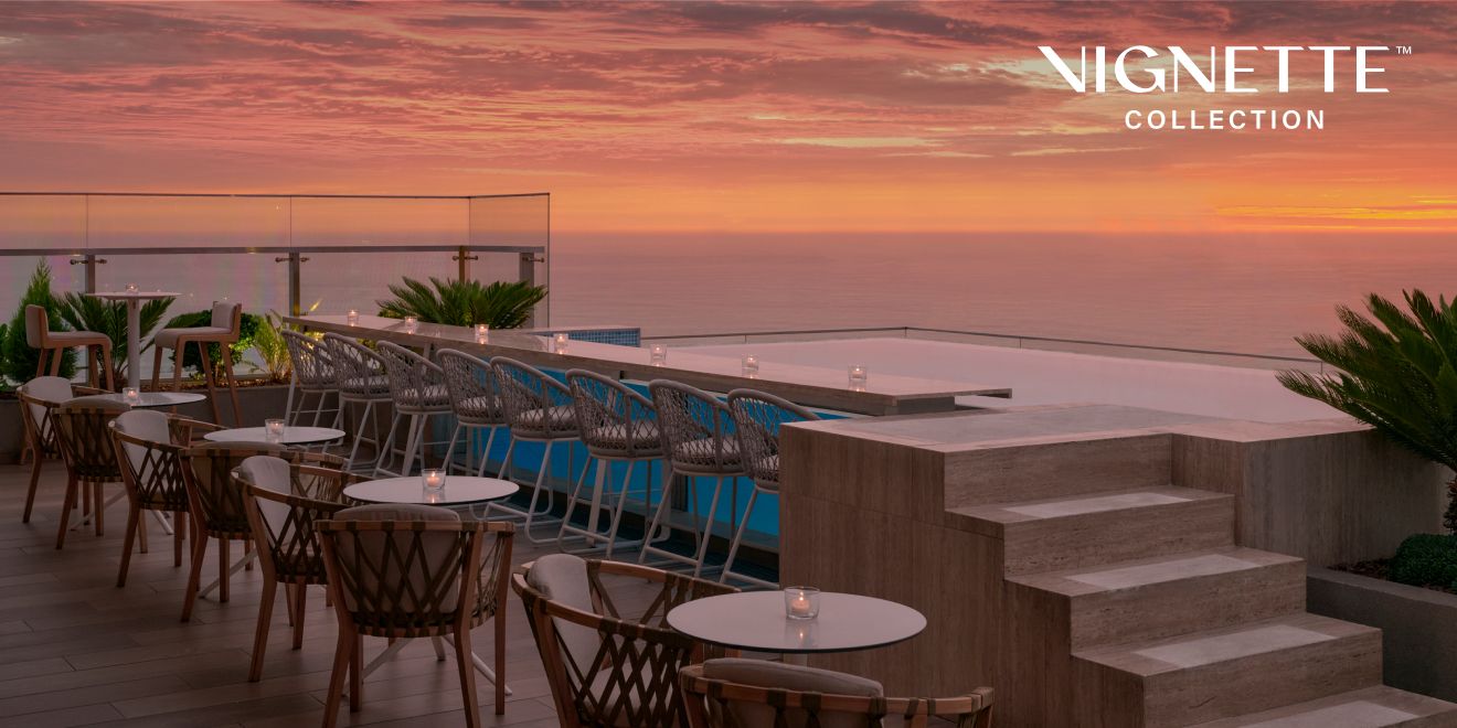 Rooftop terrace overlooking the ocean at sunset, with beautiful pink and orange sky; “Vignette Collection” logo overlaid in corner.