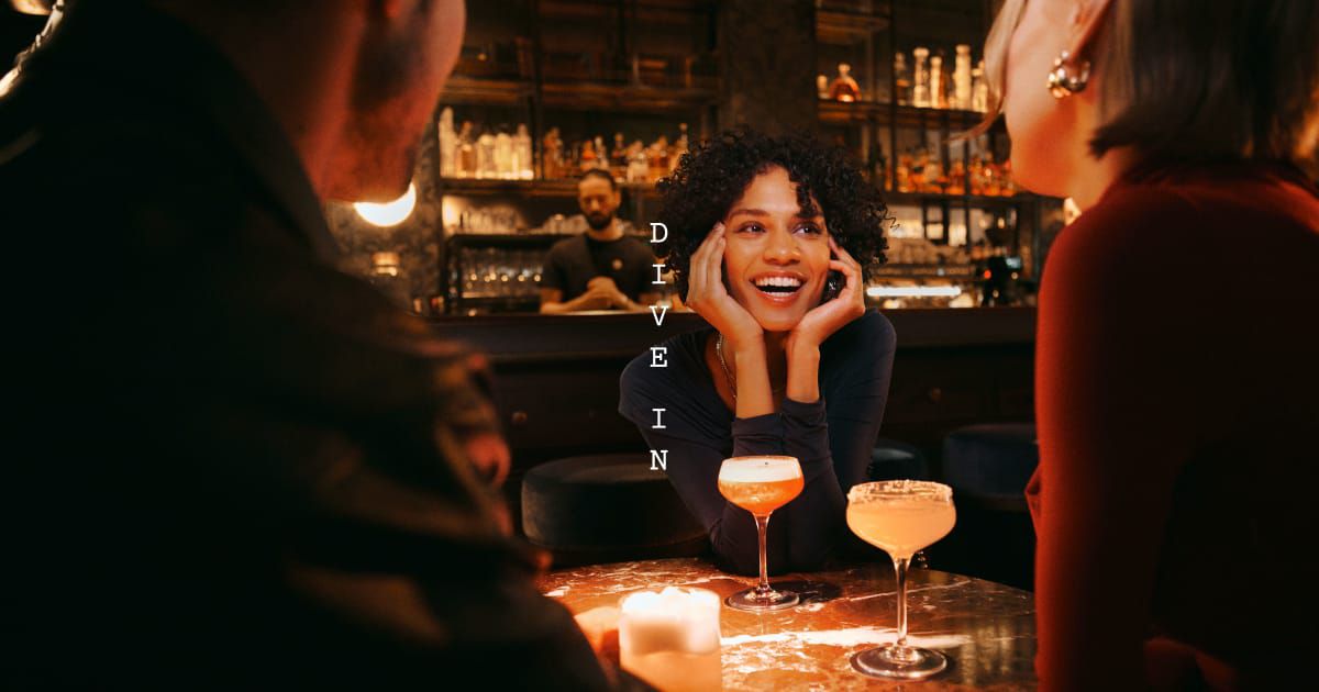 Person at a bar table chatting with two others; cocktails in foreground, bartender behind, and “DIVE IN” vertically centered.