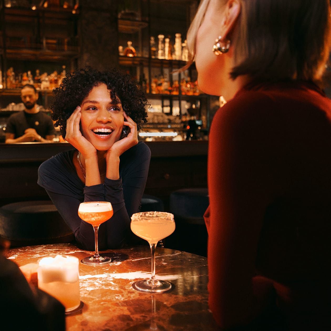 Person at a bar table chatting with two others; cocktails in foreground, bartender behind.