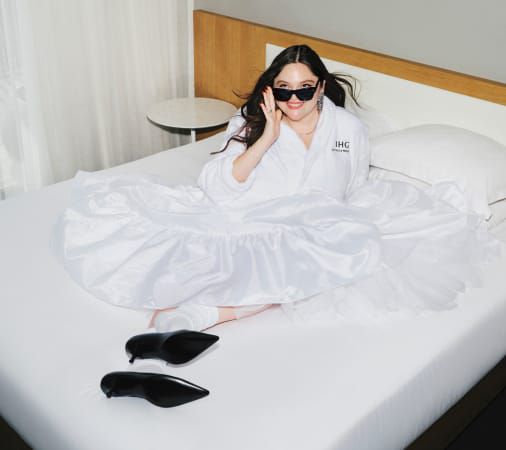 Megan Stalter sits in a hotel bed, dressed in an IHG robe, with sunglasses on and high heels kicked off.