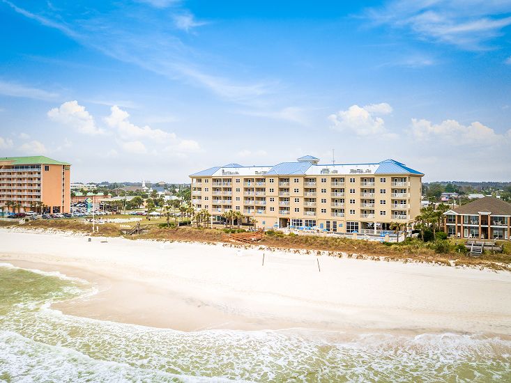 Vista aérea de un edificio de condominios frente al mar con tejados azules junto a una playa de arena blanca, con suaves olas llegando a la orilla bajo un cielo azul brillante.