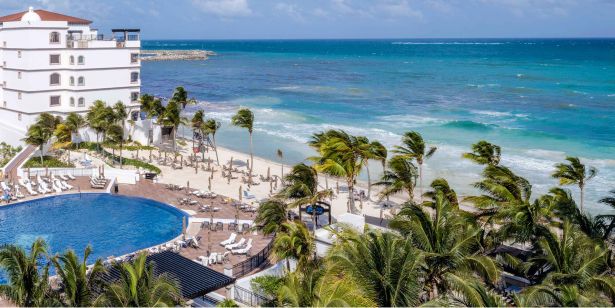 Vista aérea de un resort de playa con edificios blancos, palmeras y agua turquesa.
