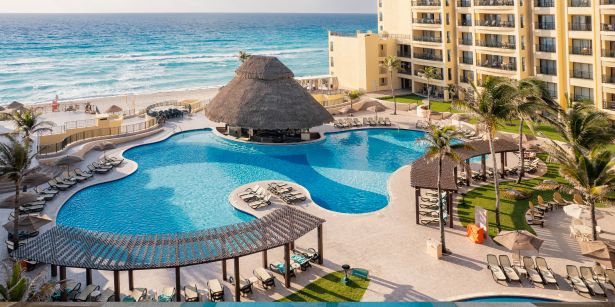 Resort frente a la playa con una piscina estilo laguna, un bar en la piscina con techo de paja y vista al mar.