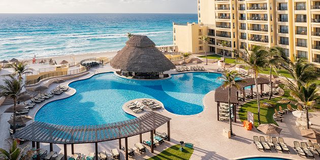 Zona de piscina del resort con un bar de techo de paja adentro, junto a un mar turquesa y un hotel de playa de varios pisos.