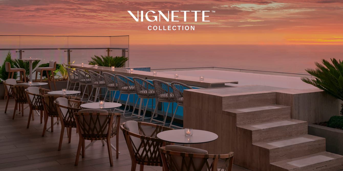 Rooftop terrace overlooking the ocean at sunset, with beautiful pink and orange sky; “Vignette Collection” logo overlaid in corner.