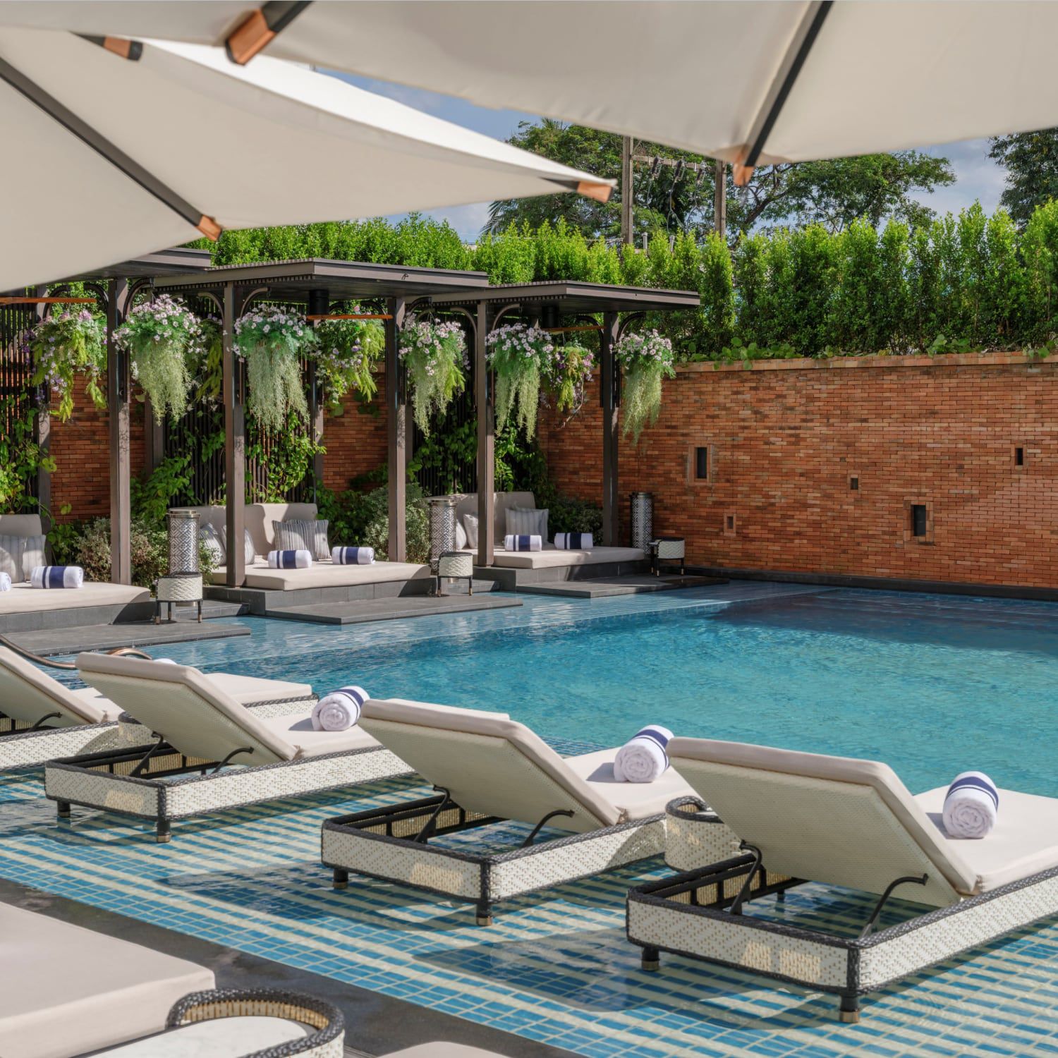 Sun loungers sit partially submerged in a pool, surrounded by hanging flower baskets, a brick wall, and green foliage.