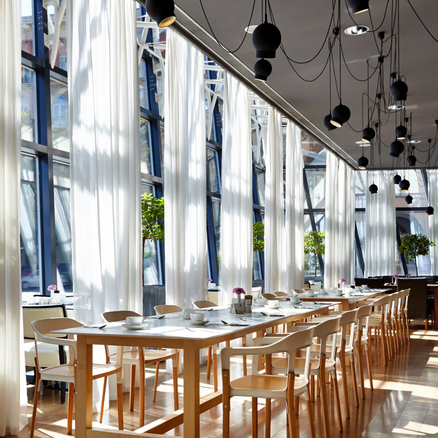 A restaurant space with floor-to-ceiling windows, gauzy curtains, and long tables set for eight.